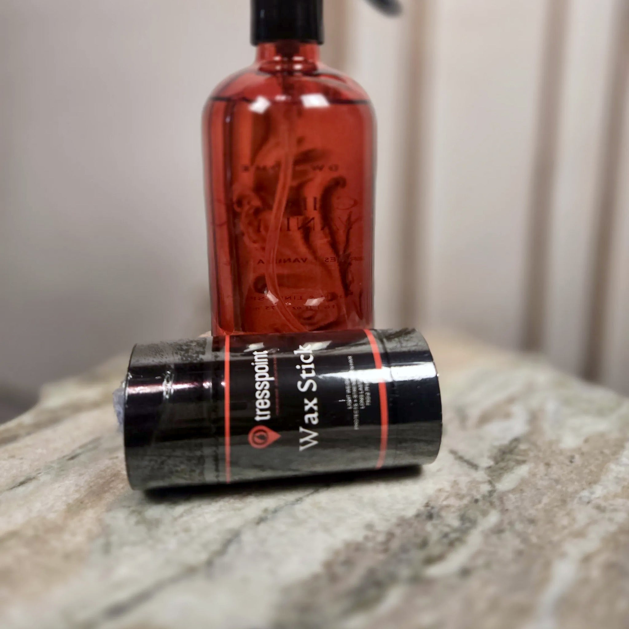 Tresspoint Wax Stick edge and strand wax, black tube with coral stripes, resting on a marble ledge beside a rose-tinted spray bottle